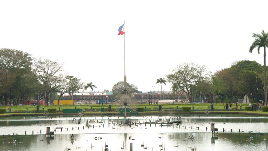 Wide shot of the Philippine flag waving over a large fountain pool in Rizal Park