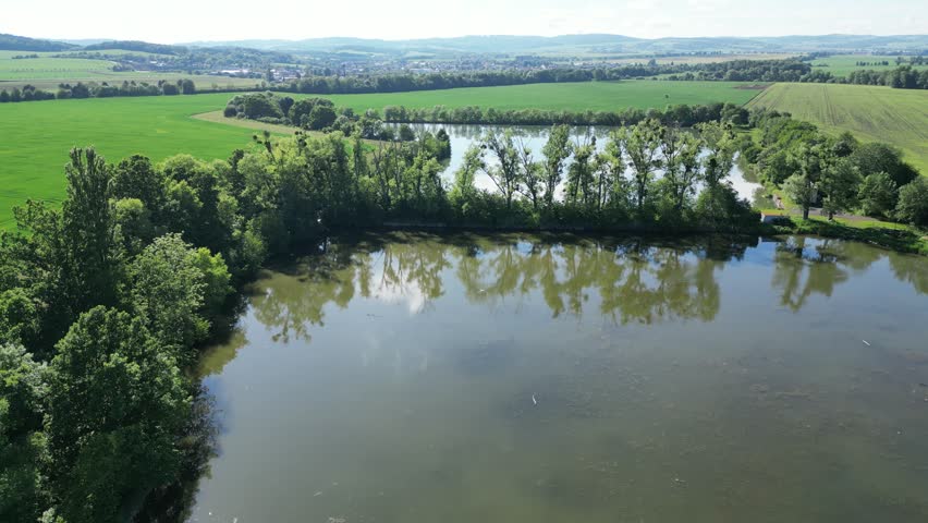 A scenic drone shot overlooking a series of tranquil ponds separated by rows of mature trees. The landscape features vibrant green agricultural fields, a clear reflection of the sky on the water surface, and a small village nestled in the rolling hills under a bright summer sun
