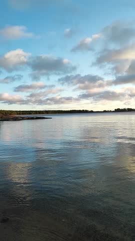 The sun sets over a tranquil body of water, casting reflections of fluffy clouds onto the surface of the still water. Clouds scatter across the sky, reflecting in the shimmering sea.