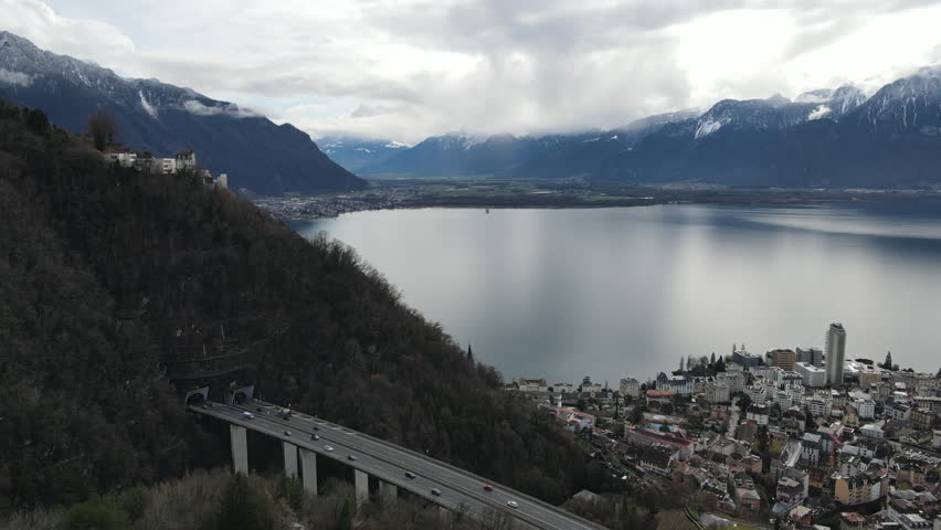 Static aerial drone shot showing a bridge and urban area of Montreux along Lake Geneva, surrounded by mountains. Clear daytime view of Swiss city infrastructure, lakeside landscape and transportation network.