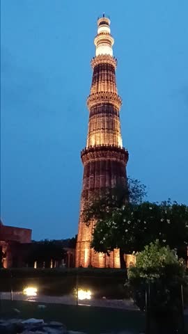 Qutub Minar in Delhi, India, illuminated at blue hour. Smooth camera pan shifts to the right after 4 seconds, showing the historic minaret from a new angle, framed by palm trees under deep blue sky.
