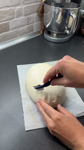A young Caucasian woman makes a cut in the shape of a wheat ear on a piece of bread with scoring lame. Homemade artisan country sourdough bread baked in home kitchen. Healthy baking, handmade food 