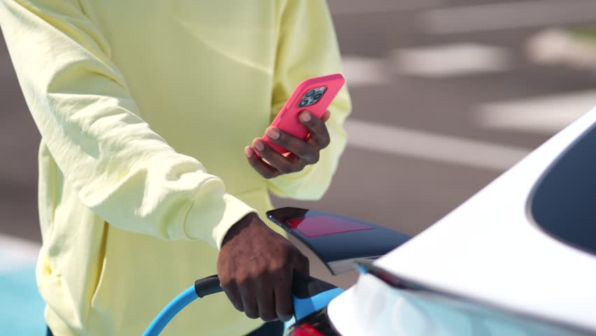 African american man using a smartphone while plugging in a charger to a white electric car