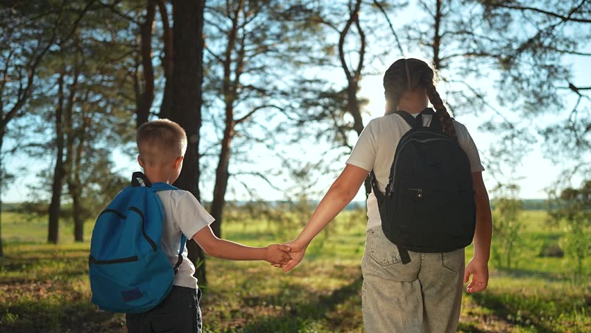 Boy walks with sister holding hand. Brother child in forest trees backpack. Girl leads boy by hand walk through forest trees. Happy child enjoy walk forest. Sister brother enjoy nature forest trees.
