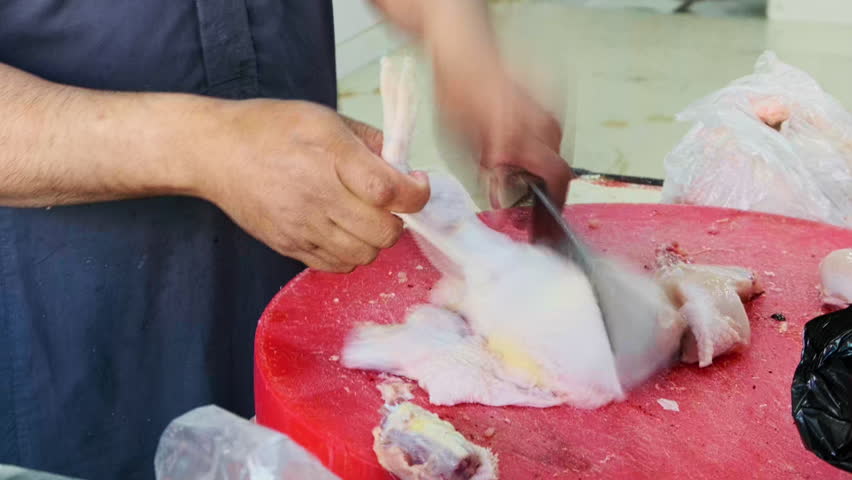 Close-up of butcher chopping raw chicken to pieces with heavy cleaver on red cutting board. One hand holds meat steady while other brings blade down.Fresh poultry processing, traditional meat handling