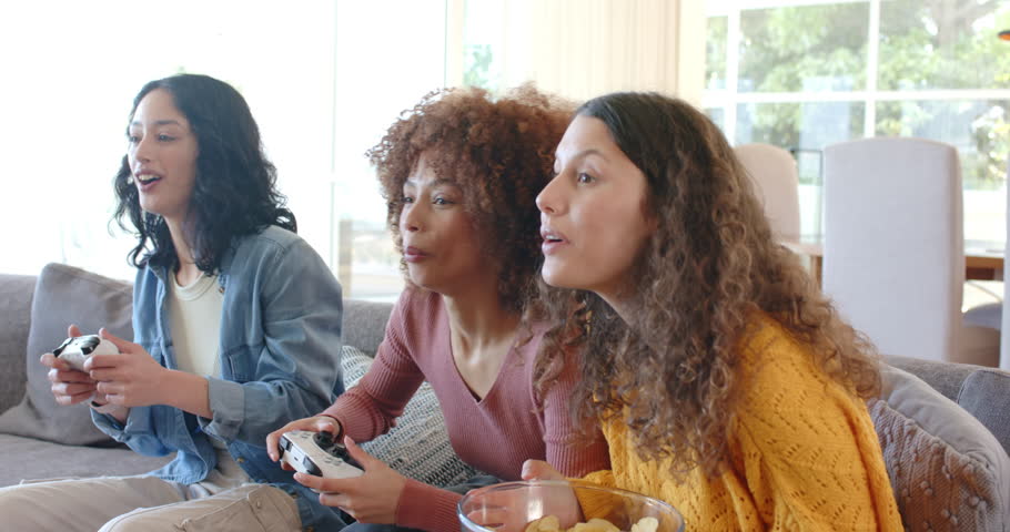 Diverse female friends gaming on sofa with controllers, chips as challenge peaking, celebrating win. Casual, friendship, engagement, vibrant, lifestyle, leisure, home