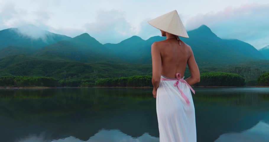 Cinematic back view of woman in white dress and conical hat looking over Tra Ly Lake, Thai Binh, Vietnam. Mirror-like water and mountain haze, perfect for tourism promos, mindfulness, and film openers