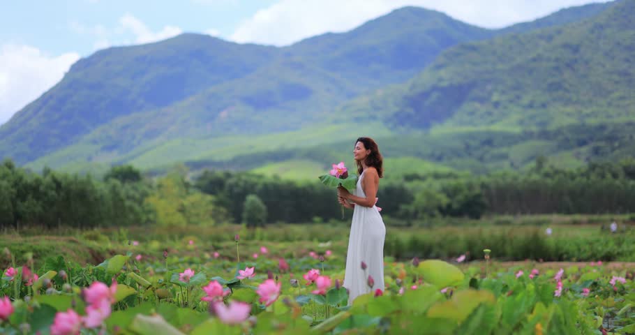 Smiling woman walks through blooming lotus and water lily pond in Tra Ly, Duy Xuyen, Quang Nam, Vietnam. Epic green mountains, perfect for tourism promos, lifestyle branding, and nature films.