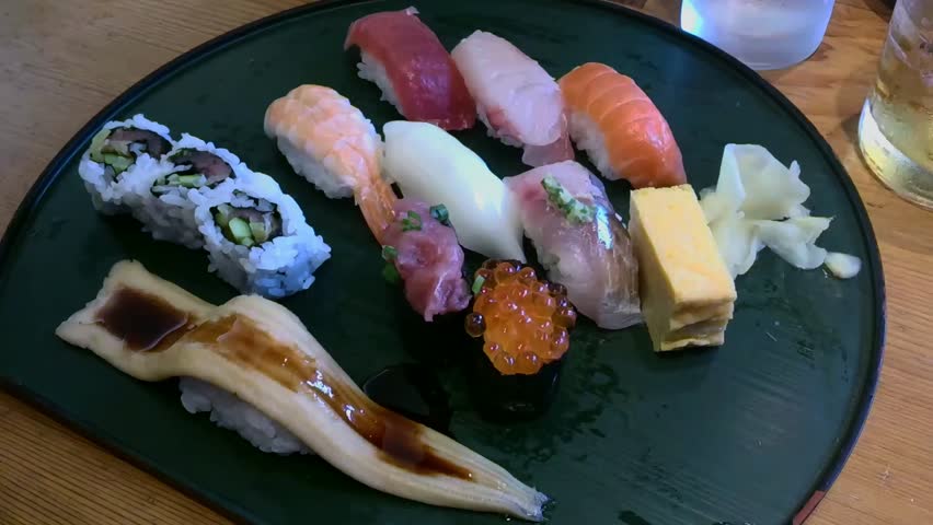 A delicious and colorful assortment of fresh Japanese sushi and sashimi platter on a black plate. Close-up view of various fresh sushi pieces including salmon, tuna, eel, shrimp, and maki rolls, served with pickled ginger on a black plate in a restaurant setting. Ideal for food blogs or menus