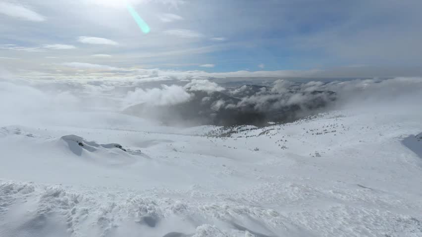 Snow-covered mountain landscape under cloudy sky with rolling clouds and distant forested slopes