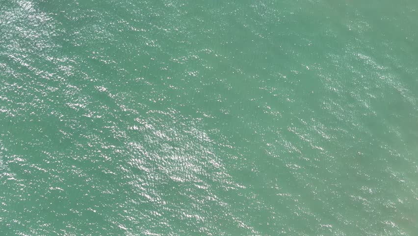 The bright sun reflects off blue ocean water showing ripples and light patterns on a summer day. Waves move gently across the surface.