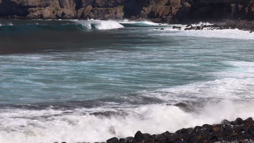 Large, foamy Atlantic waves surge toward the shoreline below the dramatic cliffs of Los Gigantes on Tenerife in the Canary Islands. Sunlight catches the white spray as the water crashes against the volcanic coast. The scene blends raw ocean energy with the iconic landscape of this well‑known holiday destination, where travelers and tourists come to experience the power and beauty of island nature.