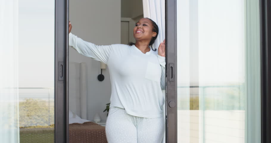 African American woman pulling curtains and opening glass door in bedroom welcoming morning light. Serenity, tranquility, brightness, natural light, lifestyle, relaxation, home