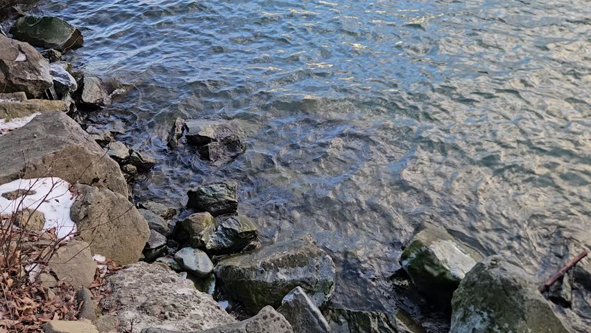 The natural landscape of Zywieckie Lake features a rocky shoreline with clear, rippling water and scattered remnants of winter snow