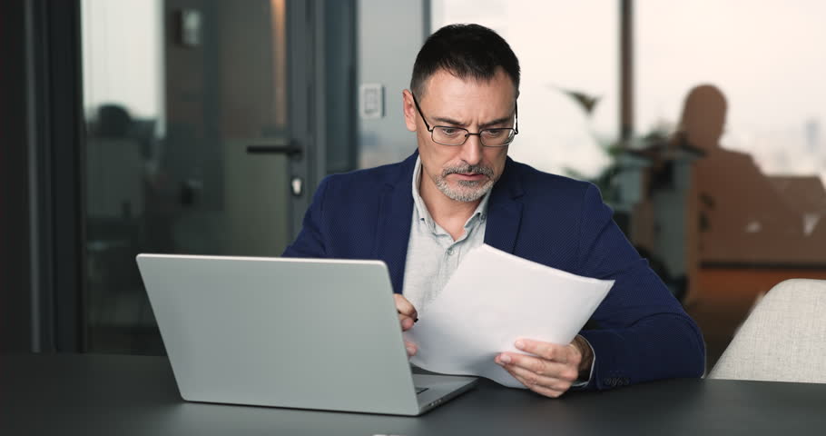 Middle-age businesman sitting at table, working on laptop, reviewing printed data, researching information, analyzing documents, preparing report, engaged in workflow in corporate office. Business