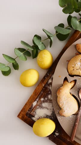 Bunny-shaped cookies are displayed on a wooden board, alongside yellow Easter eggs and greenery.