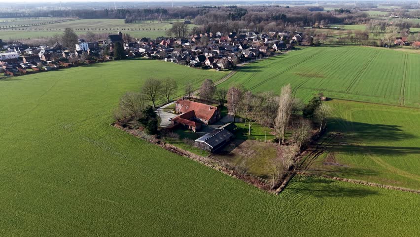 An expansive aerial drone shot of a traditional red roofed farmhouse nestled within a square of trees, surrounded by vibrant green agricultural fields and a quiet country road.