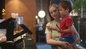 A sweet toddler boy, sitting in his mother's arms, helps her pay with a credit card at the checkout counter in a modern coffee shop. Children and money. Parents teach children how the financial system works. - Powered by Shutterstock - Get 15% off with code: PIKWIZARD15