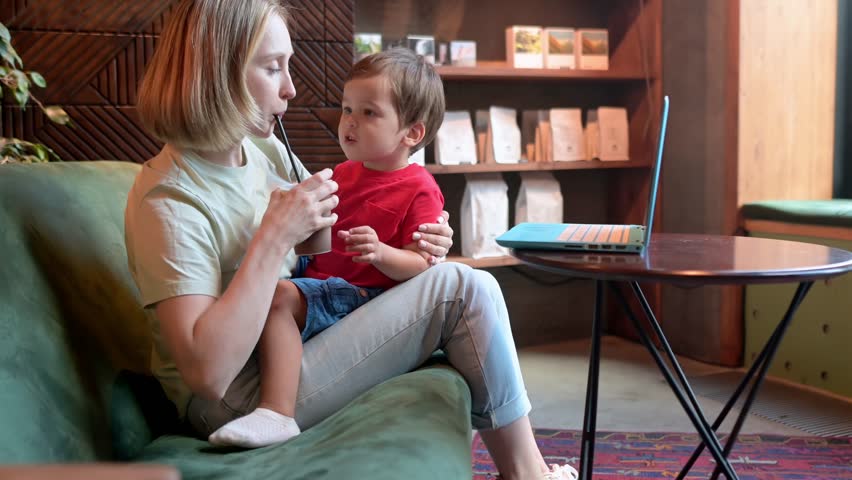 Tired mother working oh her laptop, holding her toddler son and drinking coffee sitting in a modern coffee shop. The problem of combining motherhood and work
