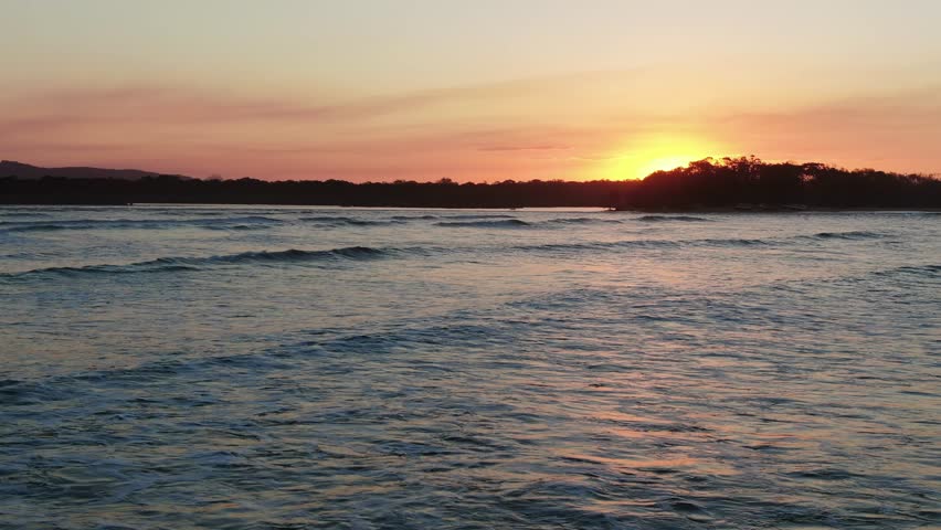 Golden sunset over gentle waves at the Noosa River mouth, Queensland, Australia. Warm evening light reflects across calm coastal water.