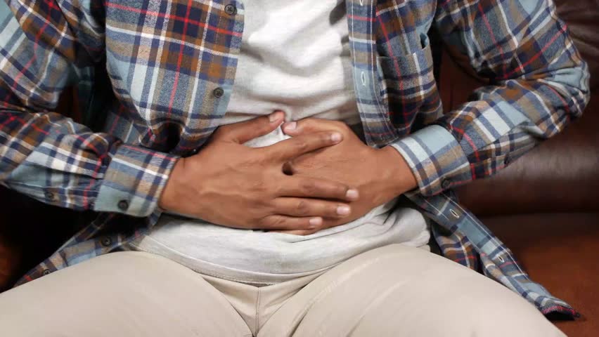 A young man experiencing abdominal pain and discomfort, holding his stomach while seated, dynamic footage capturing his struggle
