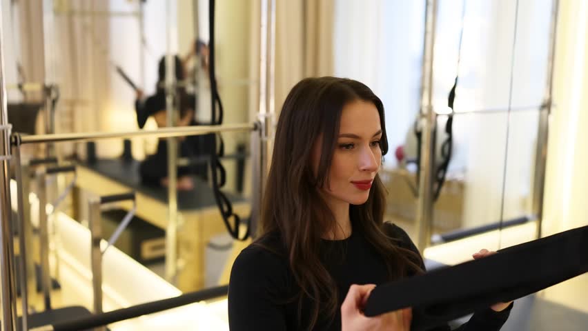 Pilates Cadillac trapeze table with wooden details, bars, and multiple handles designed for versatile training. Slim brunette model in a trendy black Pilates outfit performs controlled movements.