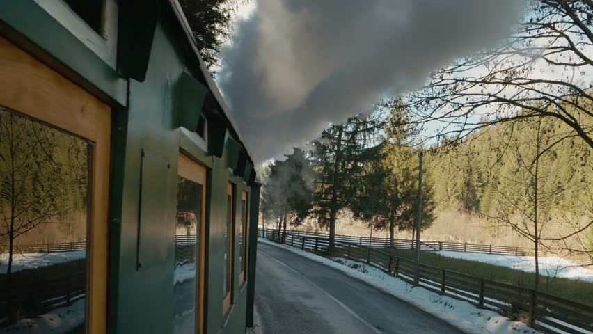 The Mocanita steam train moves through the villages of Bucovina, Romania, while it emits thick smoke. It travels past trees and snow-covered landscapes.