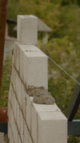 Vertical Screen: A worker skillfully lays concrete bricks to build a sturdy wall as part of house construction. The environment is a residential area with greenery.
