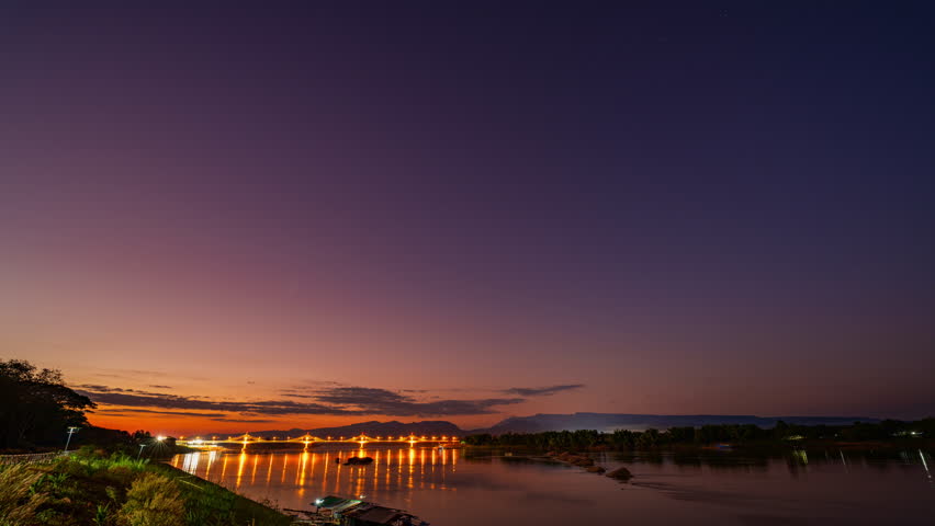 Time lapse A tranquil river scene at dusk featuring a glowing sunset sky, illuminated bridge lights, calm water reflections, and distant mountain silhouettes. peaceful landscape evokes serenity
