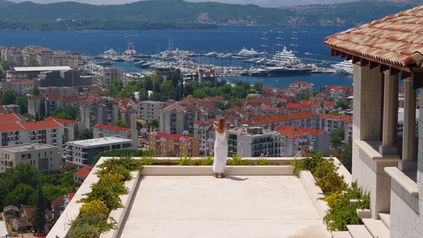 A woman in a white dress stands on a rooftop garden in Montenegro, facing a marina with superyachts and a tall masted vessel, boats moving across a deep bay.