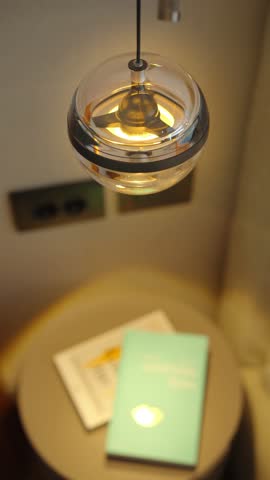 Clear glass spherical pendant light glows warm amber above a round bedside table in a Montenegro interior, two dark outlet panels on the wall, soft bokeh.