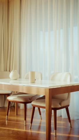 A marble topped wooden table, three upholstered chairs, vase and cup sit by sheer curtains and beige drapes in Montenegro as sunlight shifts in a slow pan.