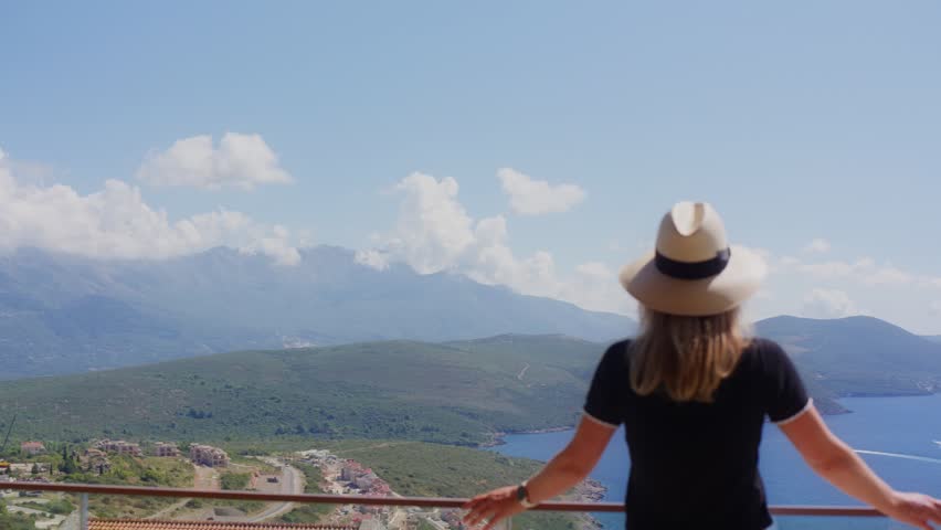 A woman in a straw hat leans on a balcony in Montenegro, midday sun. Red tiled roofs, coastal road, rounded hills, distant mountains, and a motorboat glide across the bay.