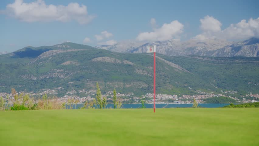 A red flagged golf hole 6 sits above a turquoise Adriatic bay, a compact town, and rocky mountains in Montenegro, midday light, wide pan with gentle breeze