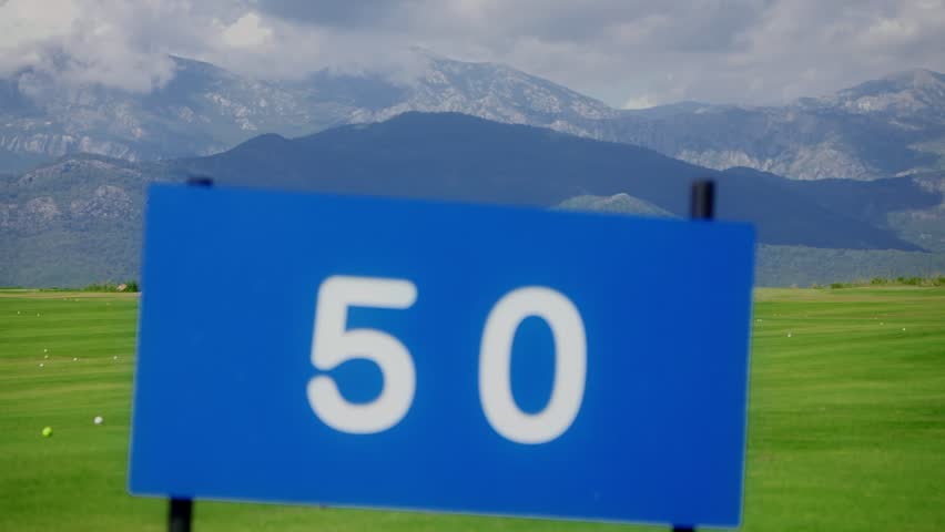 Striped fairway with 50, 100, 150 markers and practice balls, Montenegro mountains behind. Focus shifts from 50 sign to distant markers under soft daylight.