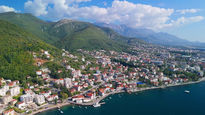 Aerial view of a Montenegro Adriatic town Tivat, curving promenade, marinas, red tiled roofs, boats and yachts, pan across harbor to steep forested hills and mountains.