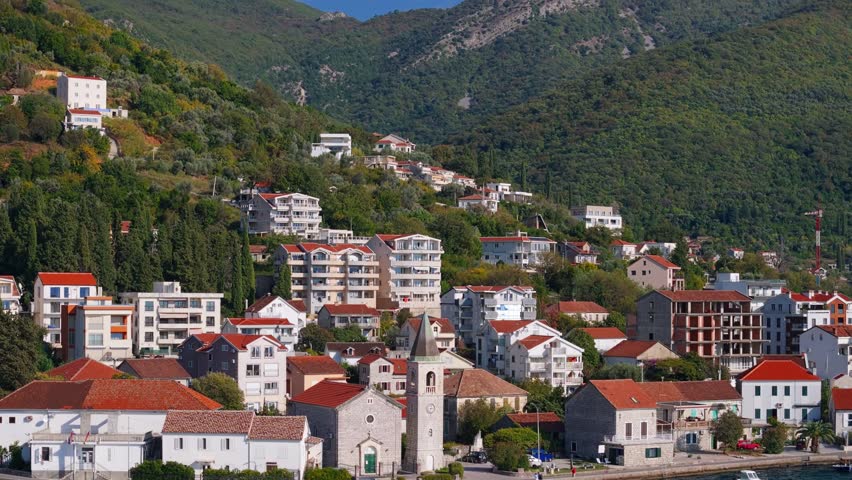Aerial view of a Montenegro coastal town with a stone church, bell tower and clock, red tiled roofs, cypress plots, and mountains, as the camera pans along the quay.