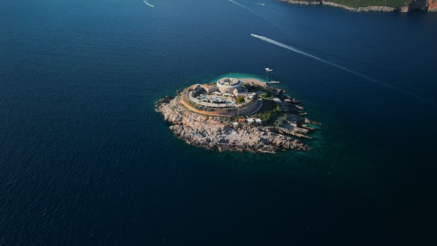 Aerial view of Mamula Island near Herceg Novi, Montenegro. The circular fortress with pools sits on rocky shores as motorboats streak wakes across deep water at midday.