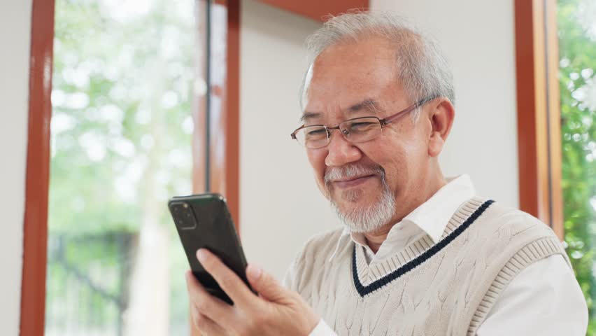 A happy Asian senior man smiles while using a smartphone at home. Digital literacy for elderly, silver economy, mobile technology, online connection, and modern retirement life.