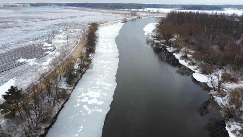 View from above:  A river flows through a snowy landscape with patches of ice along the banks. Trees line the river and fields show a mix of snow and bare soil. The setting is quiet and still.