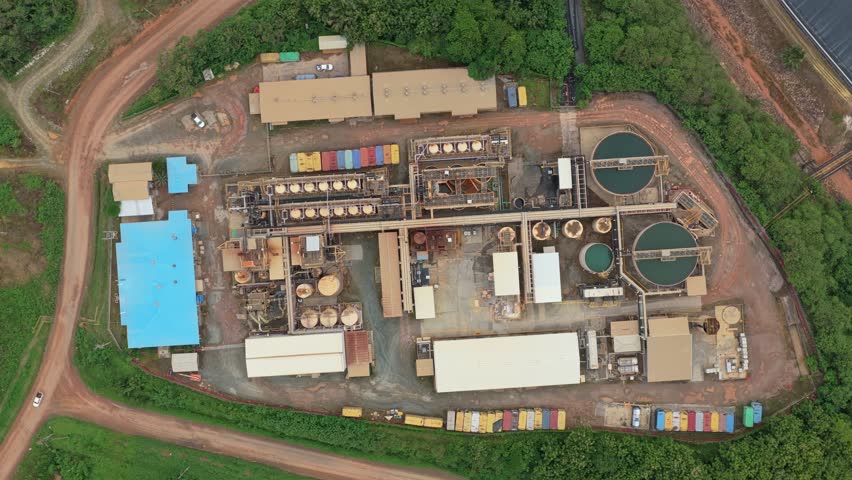 Industrial gold ore processing plant with large tanks and pipelines, filmed from a rotating aerial perspective surrounded by lush greenery