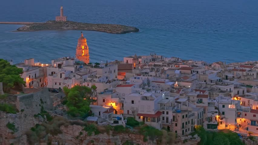 Aerial view of Vieste medieval town at night in Puglia, Italy, atmospheric resort settlement on the Mediterranean Sea with illuminated historic architecture, coastal cliffs, and dark seaside landscape