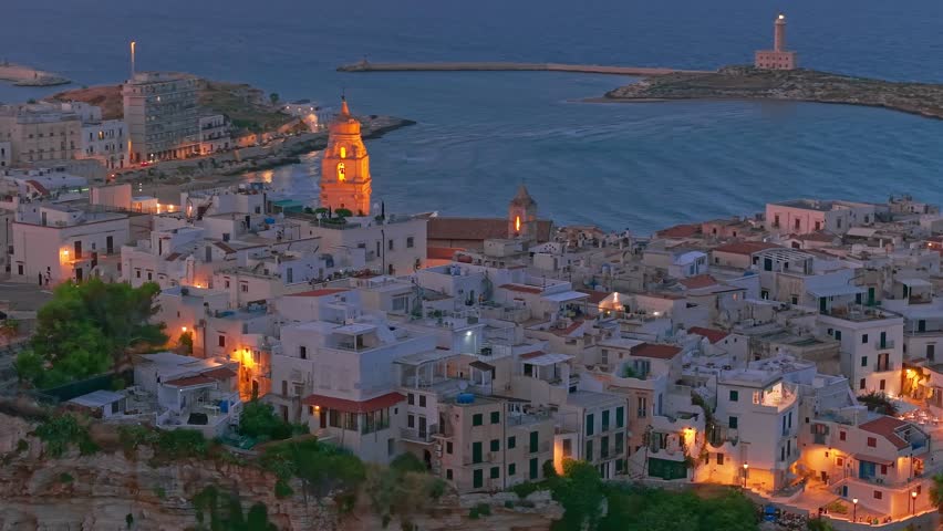 Aerial view of Vieste medieval town at night in Puglia, Italy, atmospheric resort settlement on the Mediterranean Sea with illuminated historic architecture, coastal cliffs, and dark seaside landscape
