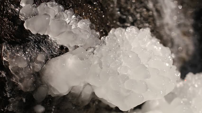 Frozen water of various shapes and textures near a waterfall. Icicles and thick layers of ice surround the cascading water, creating a dramatic winter landscape.