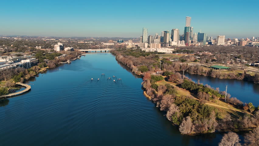 High-angle drone footage of rowing crews practicing on Lady Bird Lake in Austin, Texas. Multiple rowing shells move in formation across calm water, leaving patterned wakes behind them. Downtown Austin’s skyline rises in the distance, framed by winter trees and parkland along the shoreline. Captures outdoor fitness, collegiate rowing, teamwork, and active lifestyle in Central Texas.