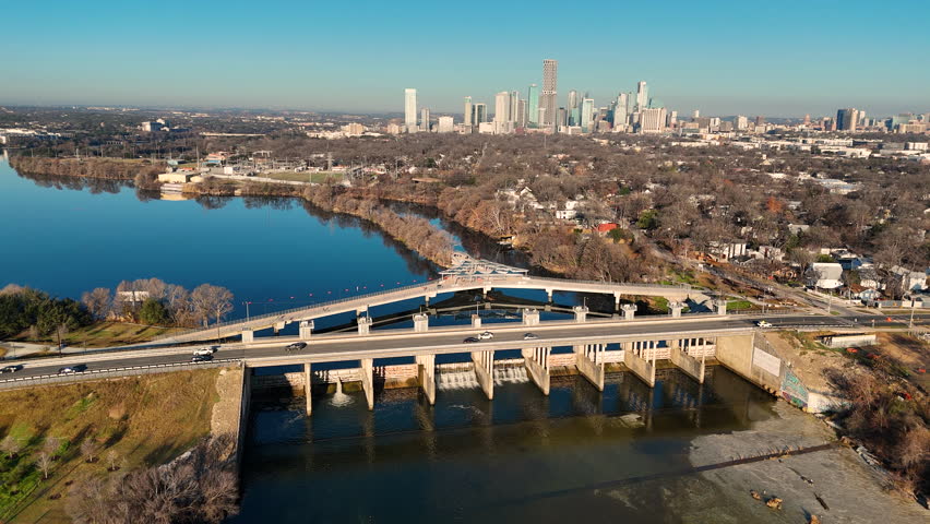 Drone footage sweeping over Longhorn Dam and the contemporary Wishbone Bridge in East Austin, Texas. The Colorado River flows beneath the pedestrian structure while still blue water fills Lady Bird Lake. Downtown Austin’s skyline stretches across the horizon under clear skies. Strong visual for infrastructure, sustainable water systems, urban planning, and Texas city growth concepts.
