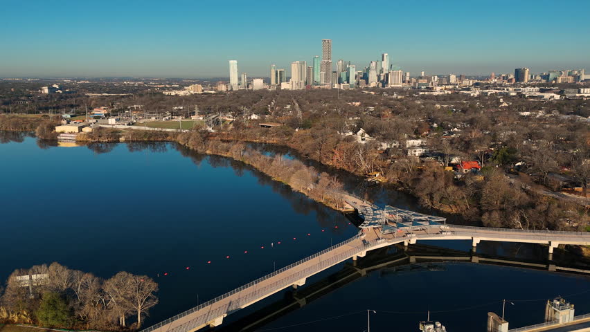 Drone footage sweeping over Longhorn Dam and the contemporary Wishbone Bridge in East Austin, Texas. The Colorado River flows beneath the pedestrian structure while still blue water fills Lady Bird Lake. Downtown Austin’s skyline stretches across the horizon under clear skies. Strong visual for infrastructure, sustainable water systems, urban planning, and Texas city growth concepts.