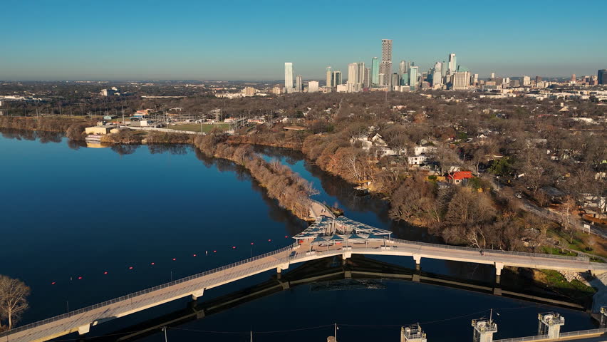 Drone footage sweeping over Longhorn Dam and the contemporary Wishbone Bridge in East Austin, Texas. The Colorado River flows beneath the pedestrian structure while still blue water fills Lady Bird Lake. Downtown Austin’s skyline stretches across the horizon under clear skies. Strong visual for infrastructure, sustainable water systems, urban planning, and Texas city growth concepts.
