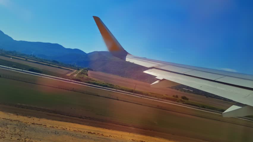 Wing of plane taking off over sunlit runway and valley, faint light with warm candle on wing, rural fields stretching to foothills, distant mountains under glowing sky, light reflected from windows