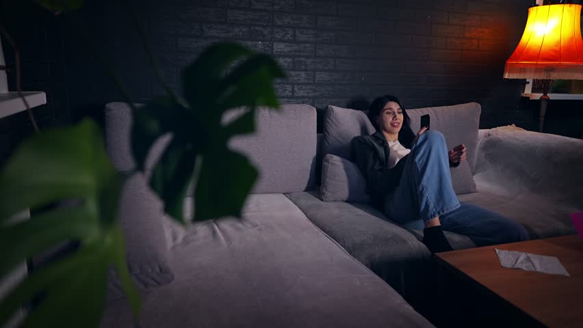 Young Caucasian woman relaxes at home on evening holding remote control and watching television. Slowmo of comfortable mood as she flips through channels. Theme of entertainment and the everyday life.